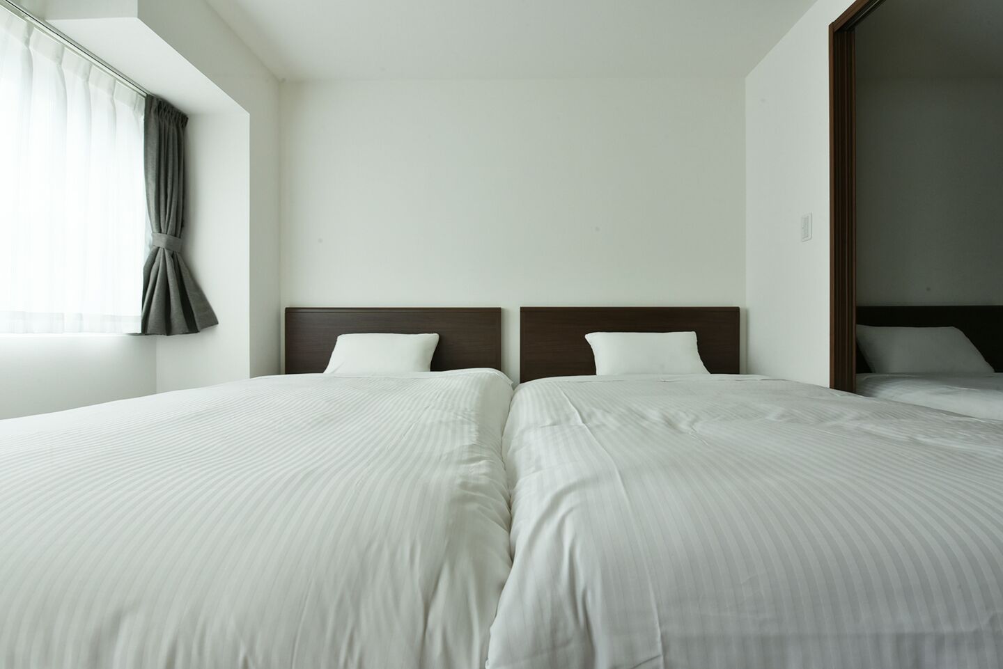 A hotel room with two beds, a large window with curtains, and a plain white wall.