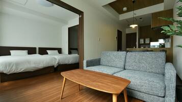 Habitació d'hotel moderna amb sòl de fusta, un sofà gris, dos llits individuals, una taula de centre de fusta i una zona de cuina visible a través d'una porta oberta.