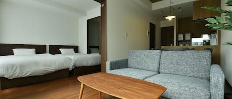 Quarto de hotel moderno com chão de madeira, sofá cinza, duas camas de solteiro, mesa de centro de madeira e cozinha visível através de uma porta aberta.
