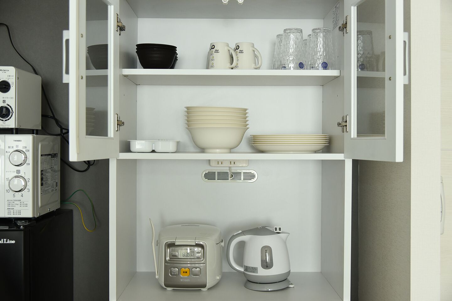 A kitchen with white cabinets, a microwave, a rice cooker, and various dishes and glasses on the shelves.
