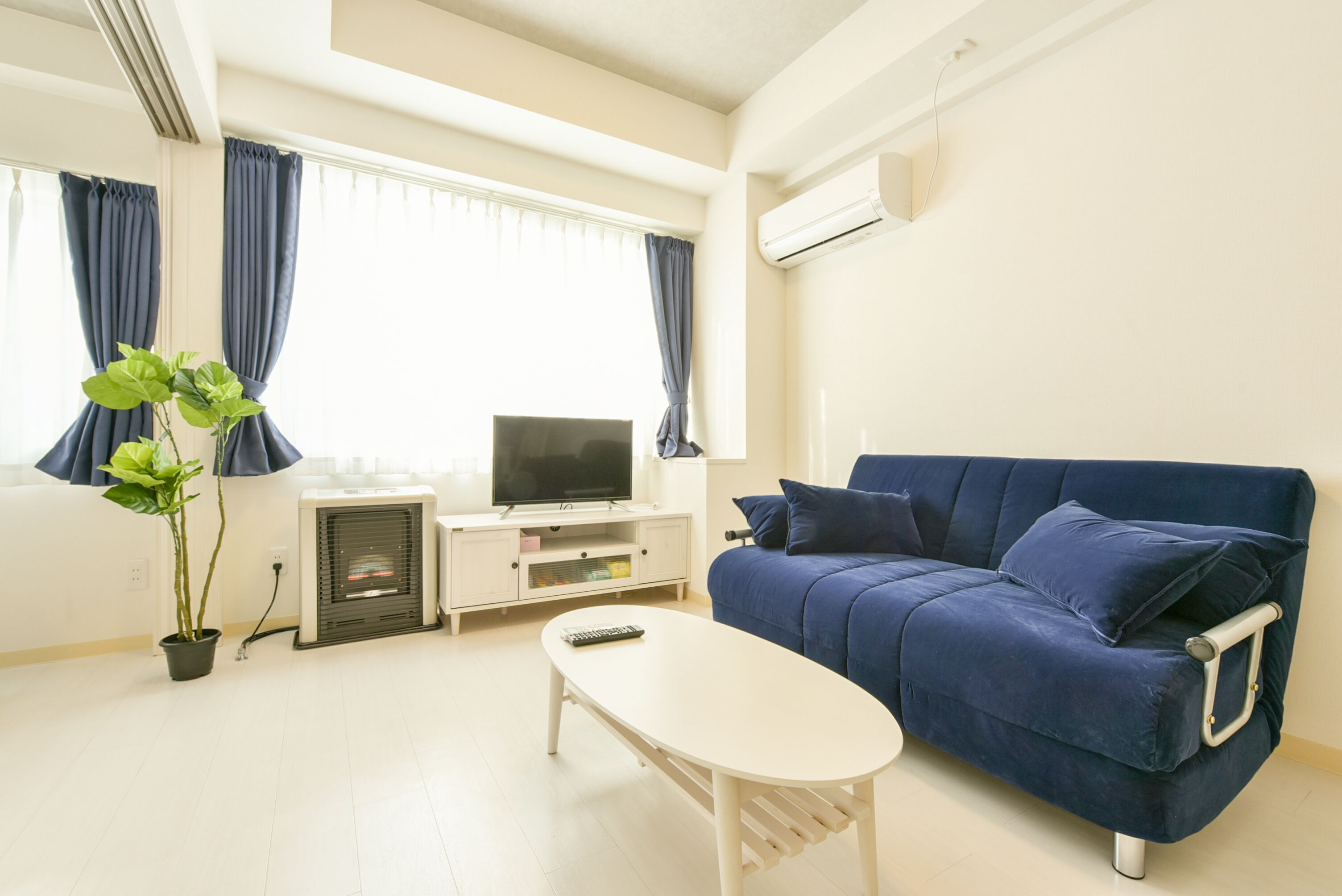 A living room with a blue sofa, a white coffee table, a TV on a white stand, a potted plant, and a window with curtains.
