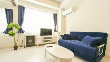 A living room with a blue sofa, a white coffee table, a TV on a white stand, a potted plant, and a window with curtains.