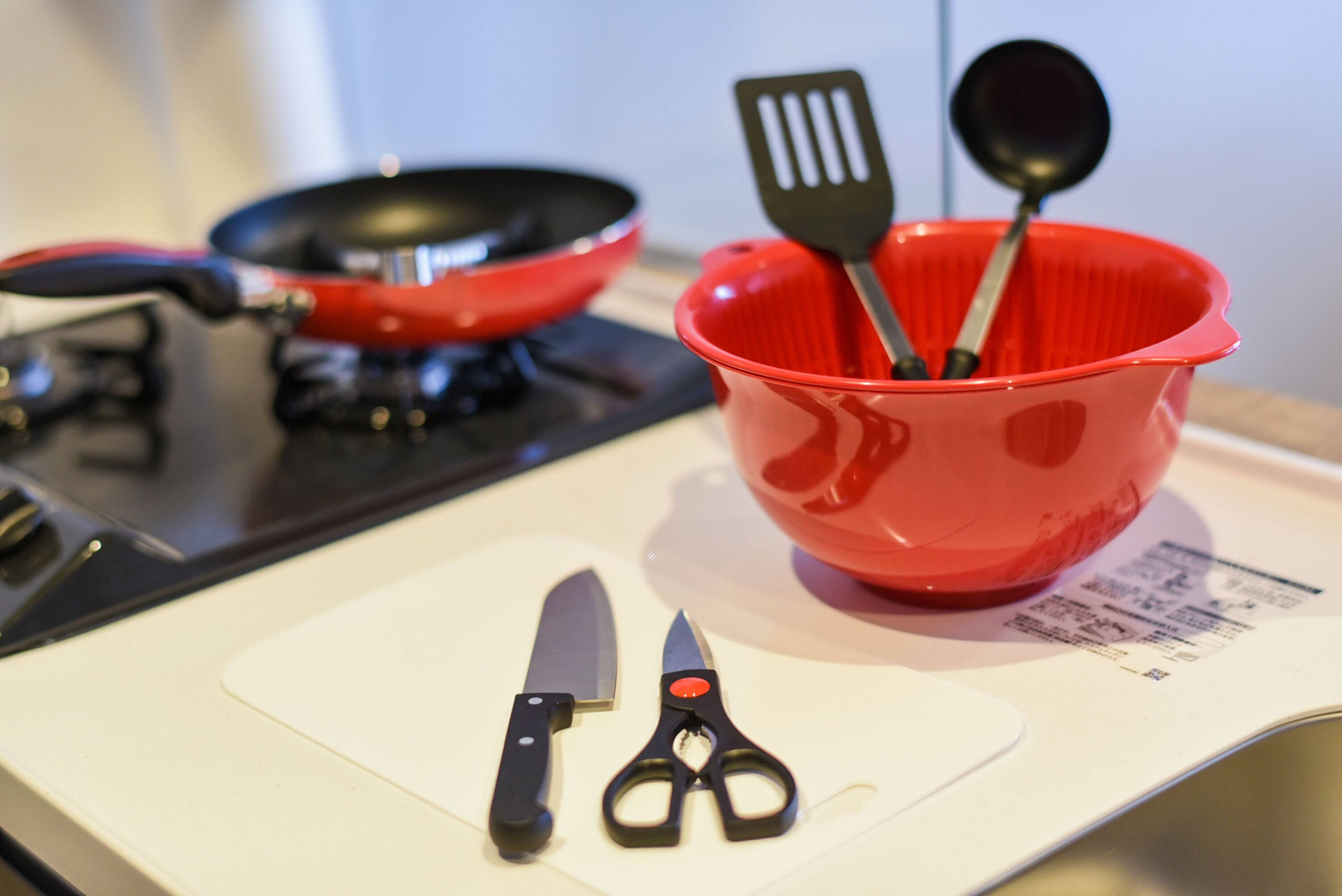 A kitchen counter with a red bowl containing a spatula and a spoon, a knife, and scissors.