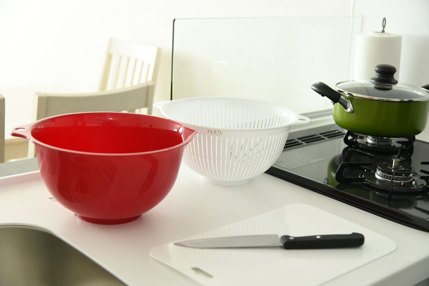 Un plan de travail de cuisine avec un saladier rouge, une passoire blanche, une casserole verte et un couteau sur une planche à découper blanche.
