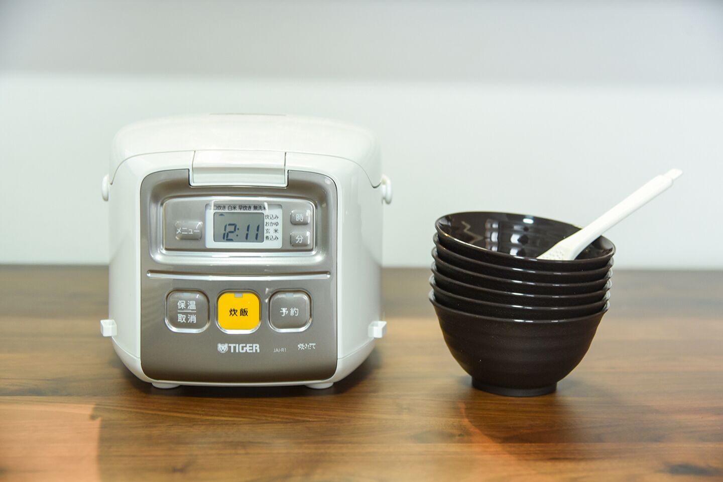Uma panela de arroz elétrica branca com visor digital e botões, ao lado de uma pilha de tigelas pretas e uma colher branca.