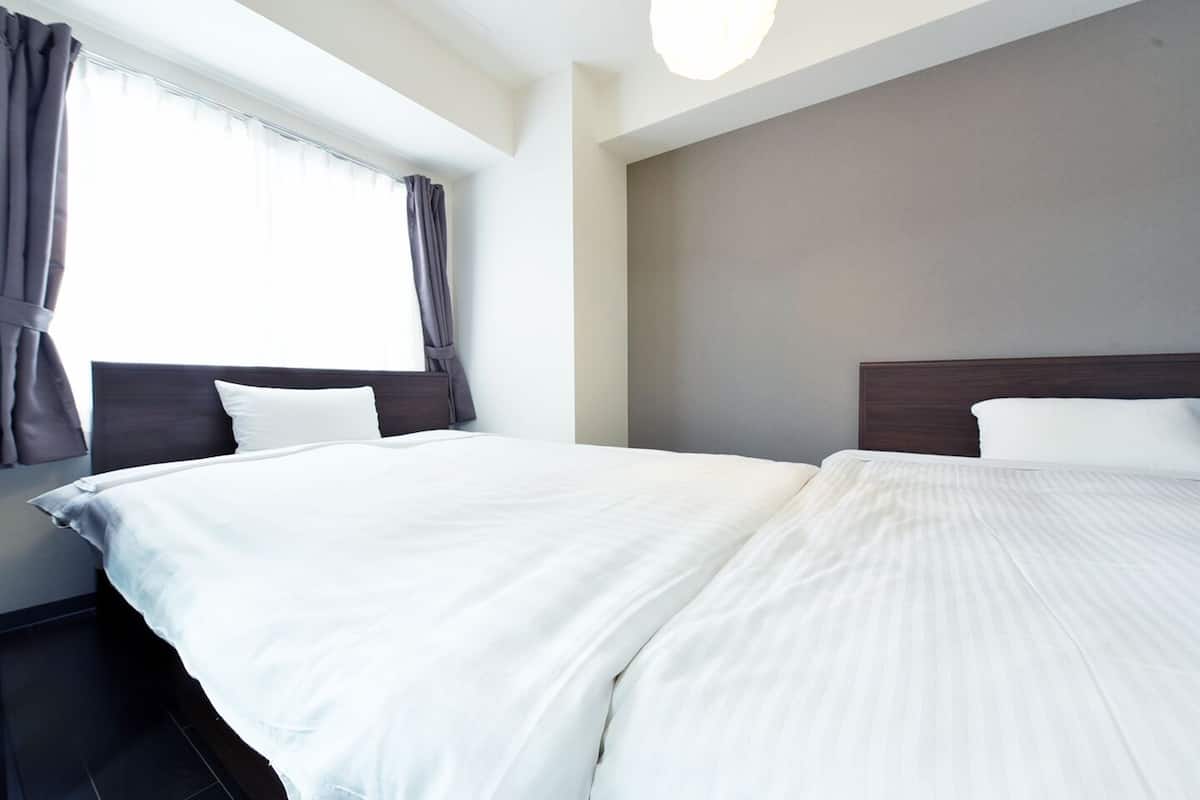 Habitación de hotel con dos camas, un ventanal con cortinas y una paleta de colores neutros.