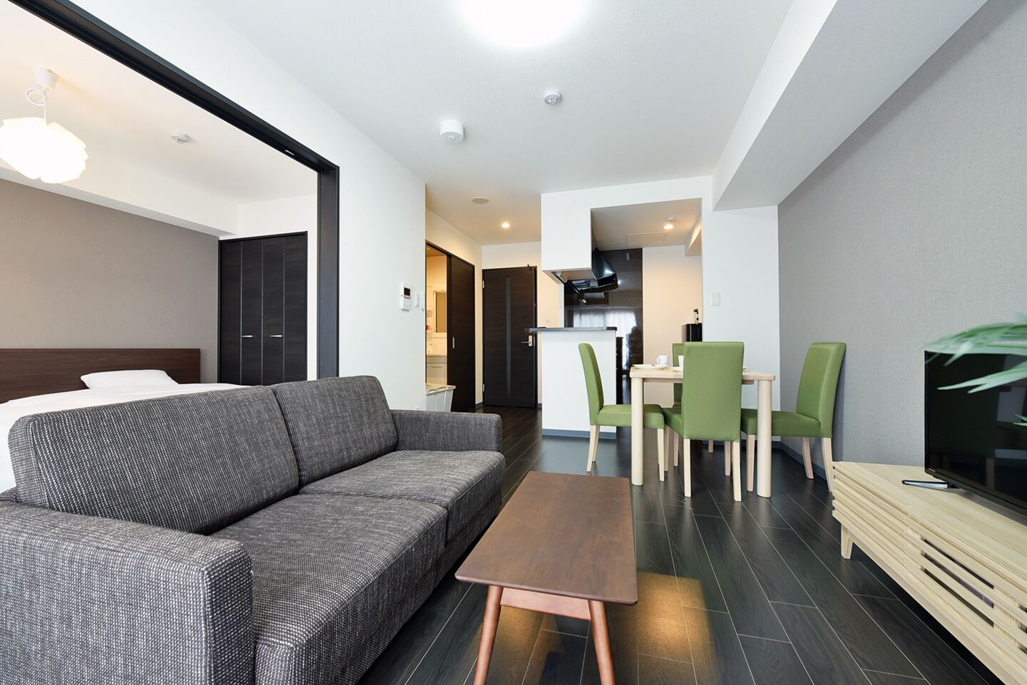 A modern living room with a grey sofa, a wooden coffee table, and a TV on a wooden stand. There is a dining area with green chairs and a kitchen in the background.