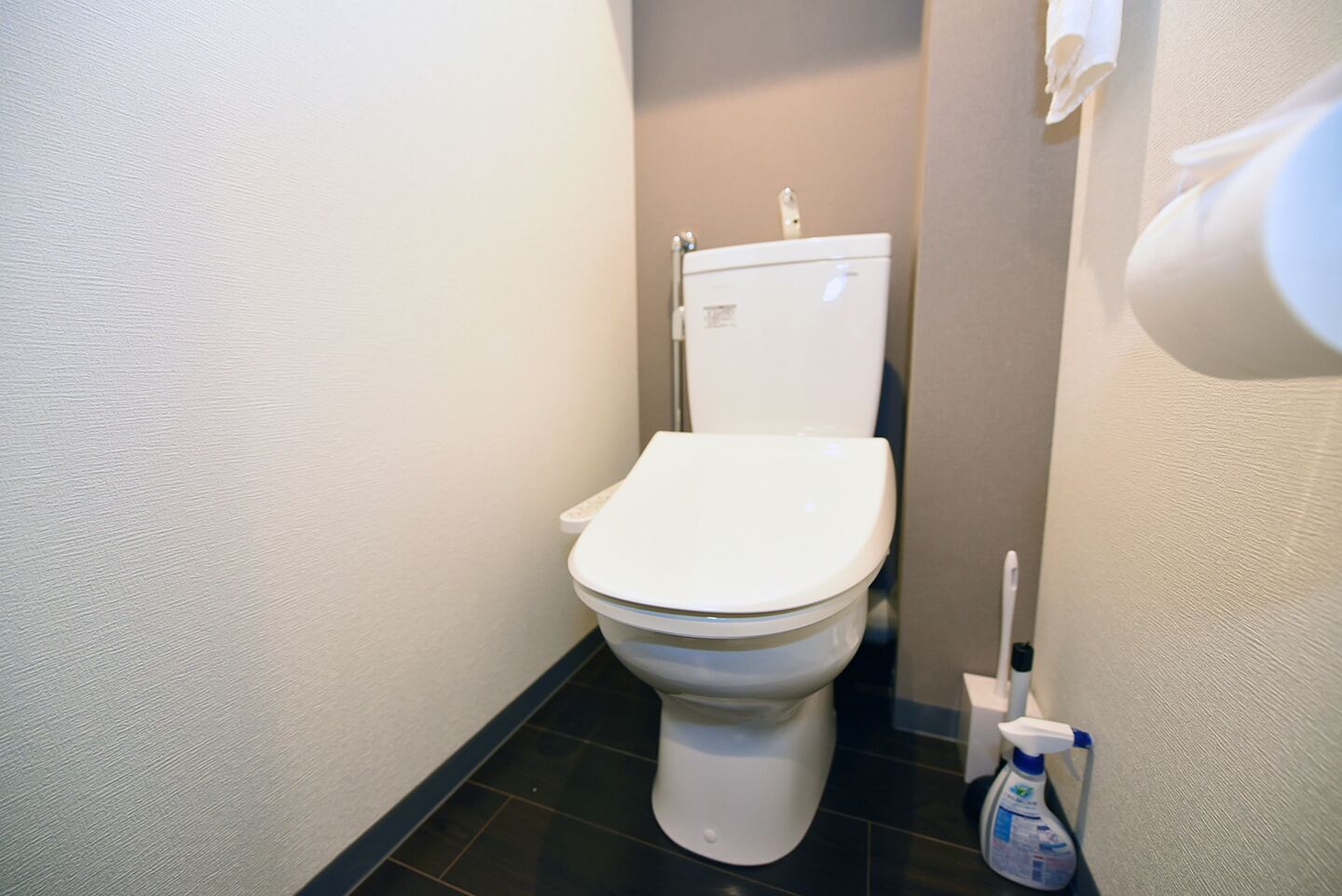 Un inodoro blanco en un baño con azulejos negros, un dispensador de toallas blanco y una botella de limpiador en el suelo.