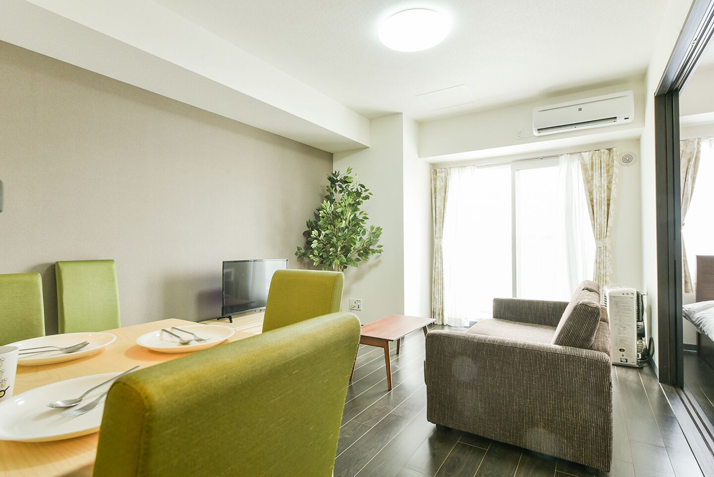 Habitación de hotel con comedor, sofá, televisión y un amplio ventanal con cortinas.