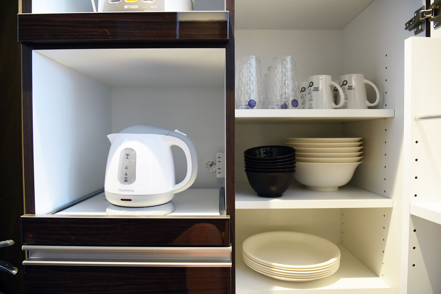 A kitchen with a white kettle on the left and shelves with dishes and cups on the right.