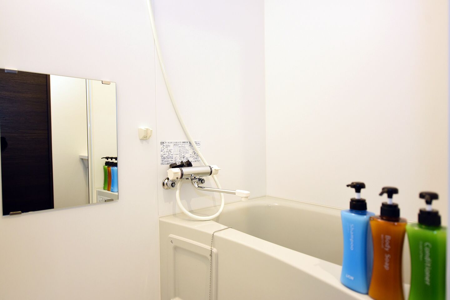 Een badkamer met een bad, een douche en een spiegel. Drie shampoo flessen staan op de rand van het bad.