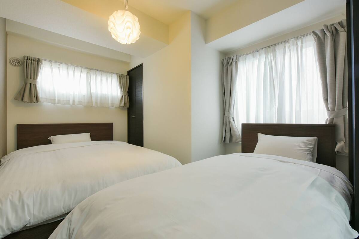 Habitación de hotel con dos camas individuales, una lámpara moderna y ventanas grandes con cortinas.