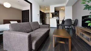 A modern hotel room with a sofa, a wooden table, a TV, and a dining area with chairs. - Sun Court Hongodori Garden Hills (Sapporo)