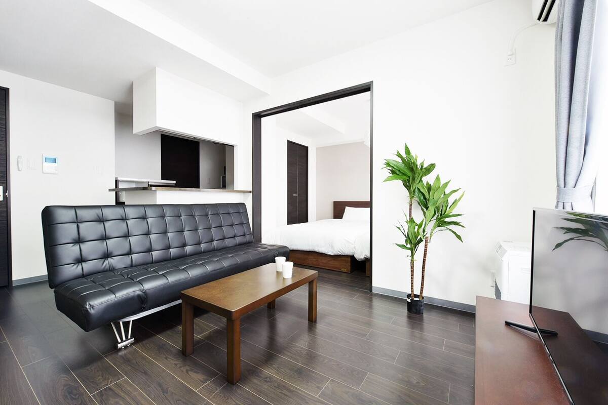 Ein modernes Wohnzimmer mit einer schwarzen Ledersofa, einem Holztisch und einem Bett im Hintergrund. Zwei Kübelpflanzen und ein Flachbildfernseher auf einem Standfuß sind ebenfalls zu sehen.