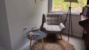 Eine gemütliche Ecke mit einem gepolsterten Sessel, einem kleinen runden Tisch mit einer Zeitschrift, einer Stehlampe und Blick ins Grüne durch ein Fenster.
