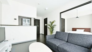 Een moderne hotelkamer met een grijze bank, een witte tv-meubel, een bed met houten hoofdeinde, en een grote potplant.