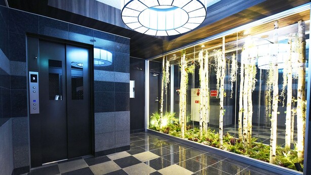 Een lift met een digitaal display en noodknoppen, een zwart-wit vloer en een glazen wand met verticale tuinen.