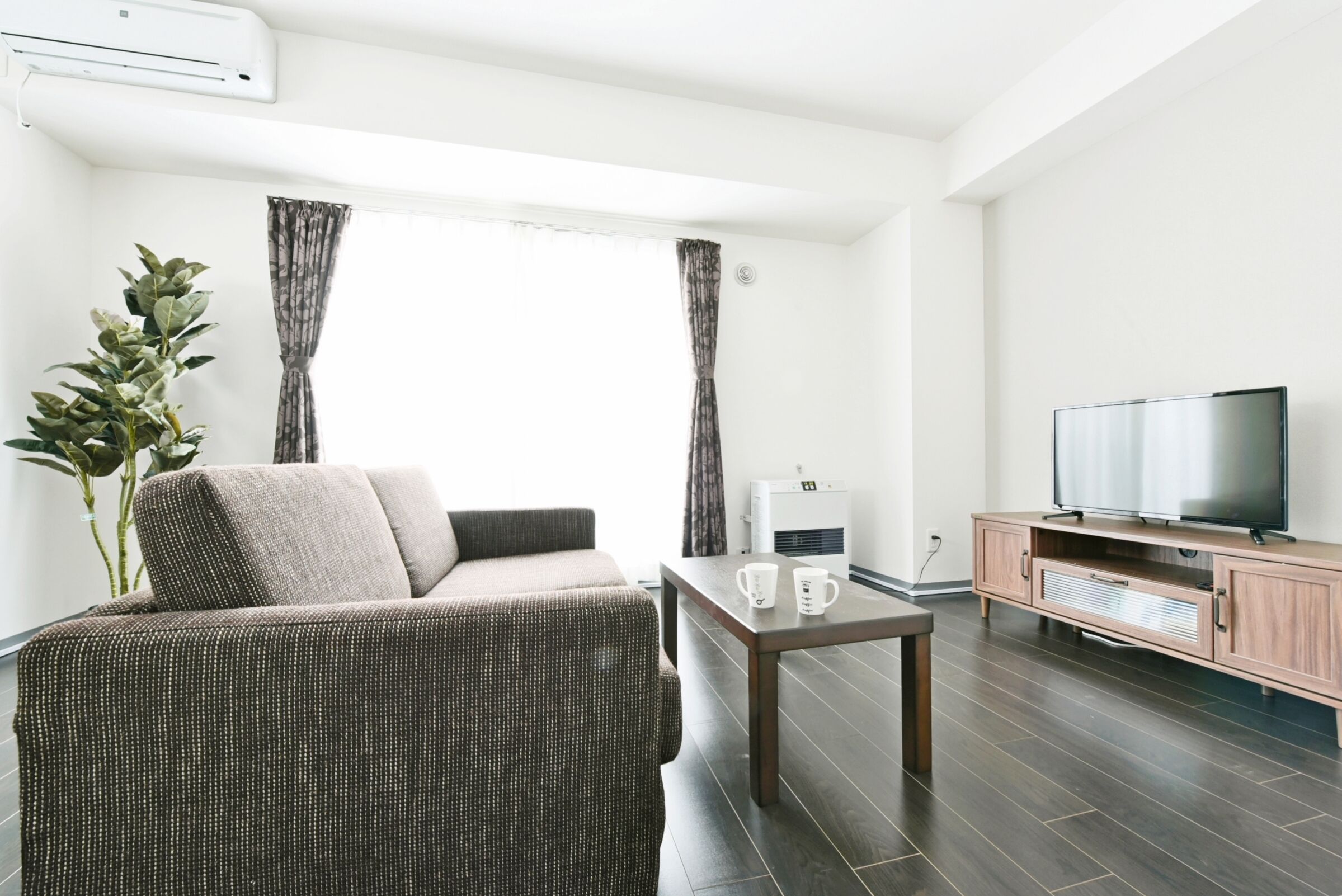 A modern living room with a sofa, a coffee table, a TV on a wooden cabinet, and a large window with curtains.