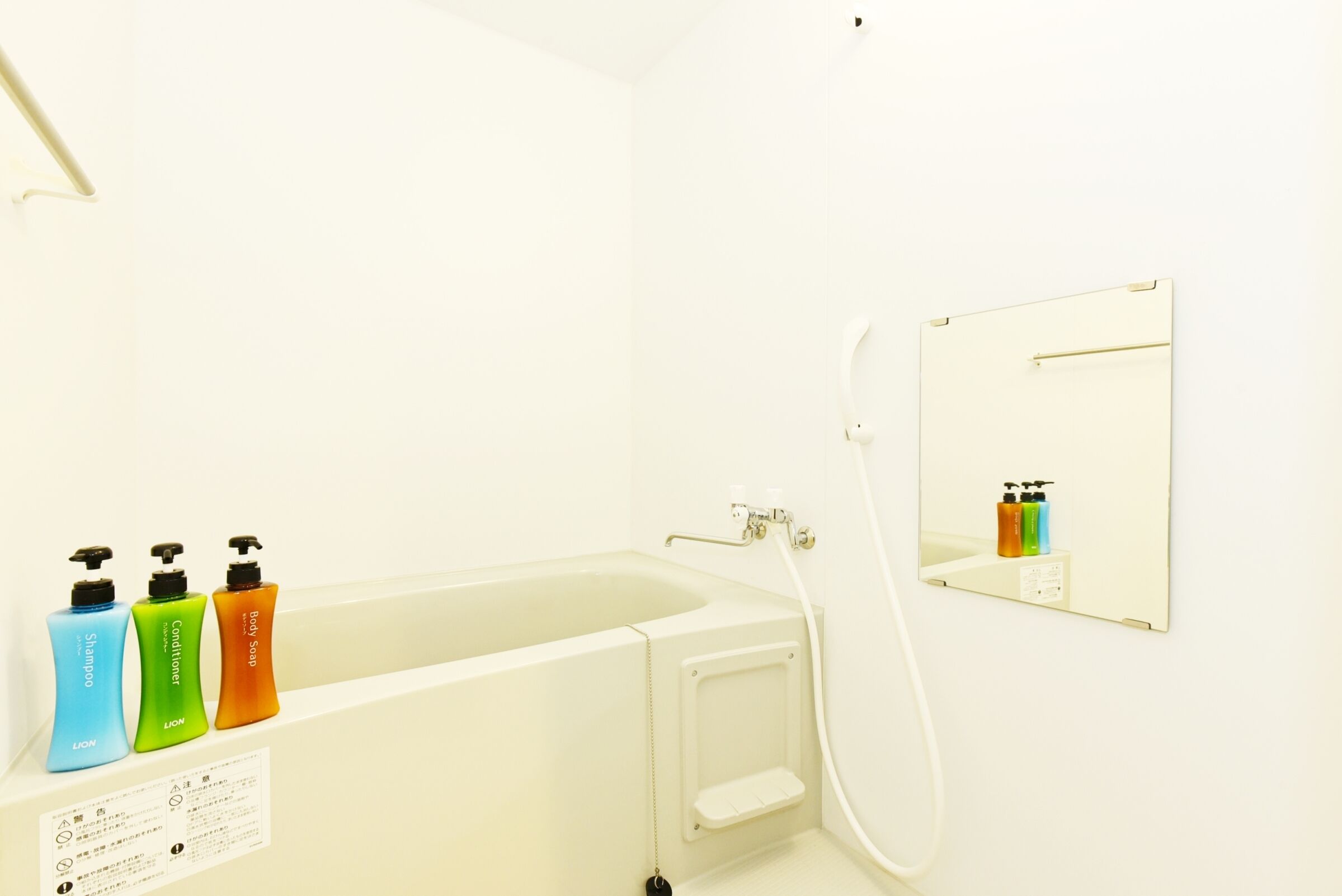 Une salle de bain avec une baignoire, une douche et un miroir. Trois bouteilles de shampoing, d’après-shampoing et de gel douche sont posées sur le bord de la baignoire.