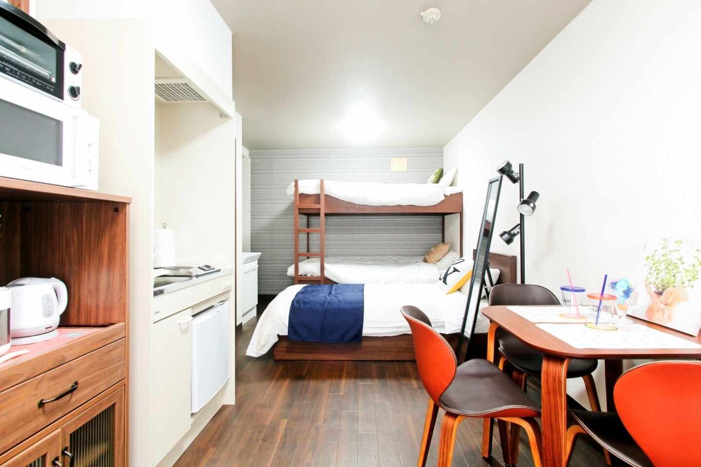 Une chambre d’hôtel compacte comprenant un lit superposé, une kitchenette, un bureau avec des chaises et une petite table avec une lampe.