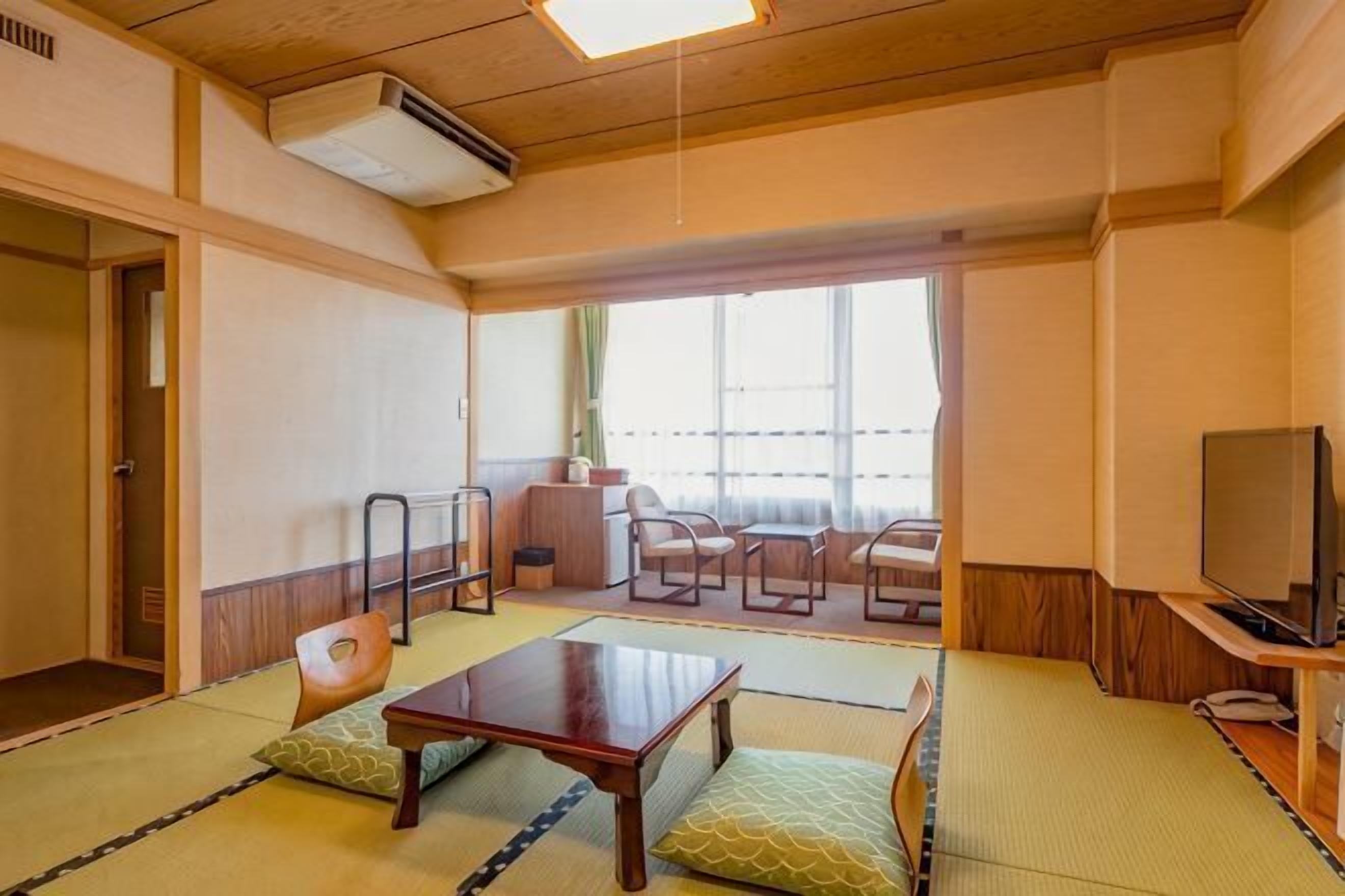 A traditional Japanese room with tatami flooring, a low table, cushions, a TV, and a view through a large window.