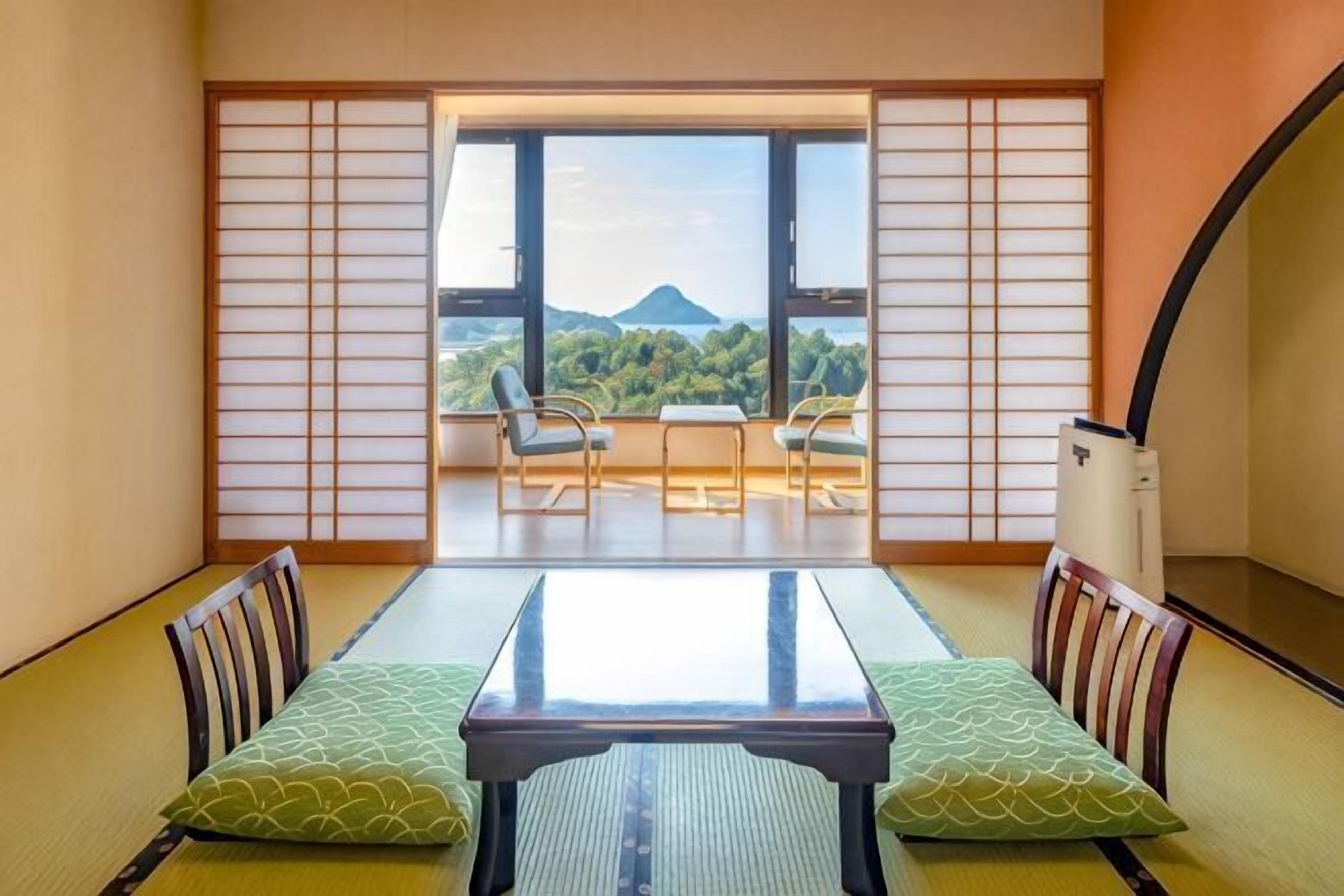 A traditional Japanese room with tatami flooring, a low table with cushions, and a sliding door leading to a balcony with a view of a mountain.