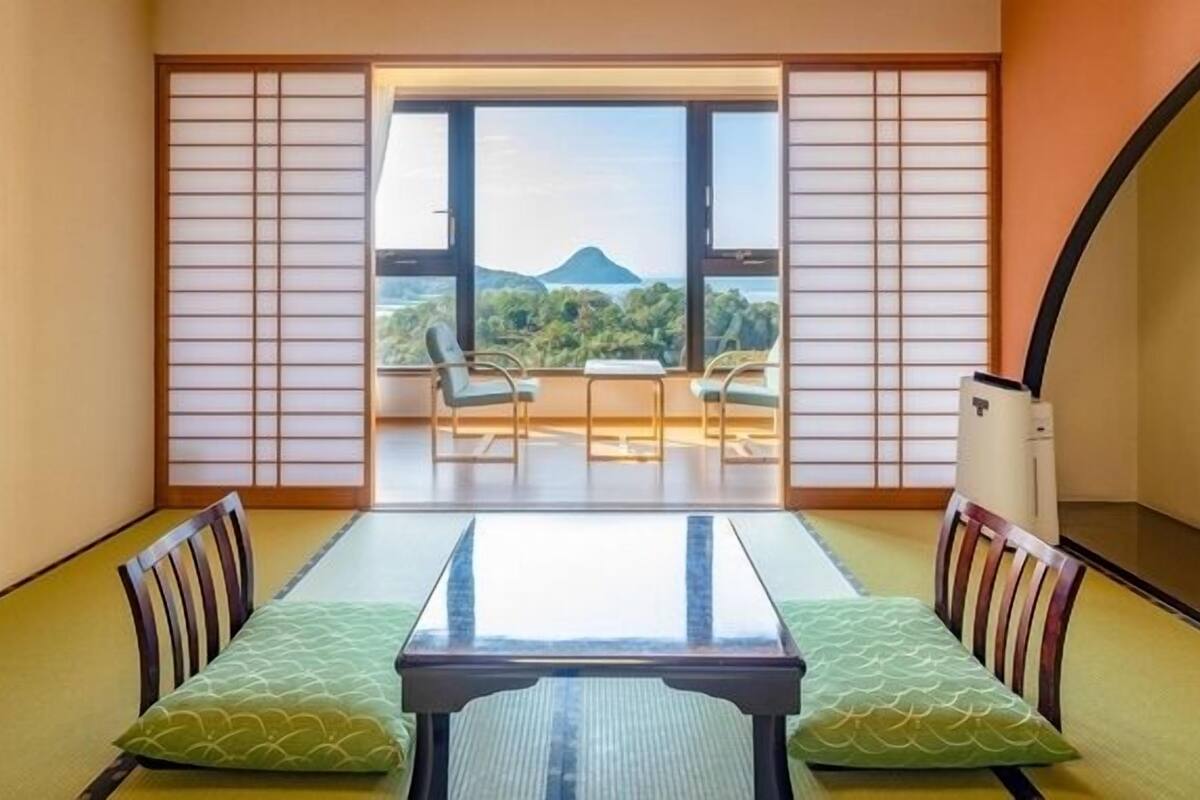 A traditional Japanese room with tatami flooring, a low table with cushions, and a sliding shoji screen leading to a balcony with a view of a mountain.