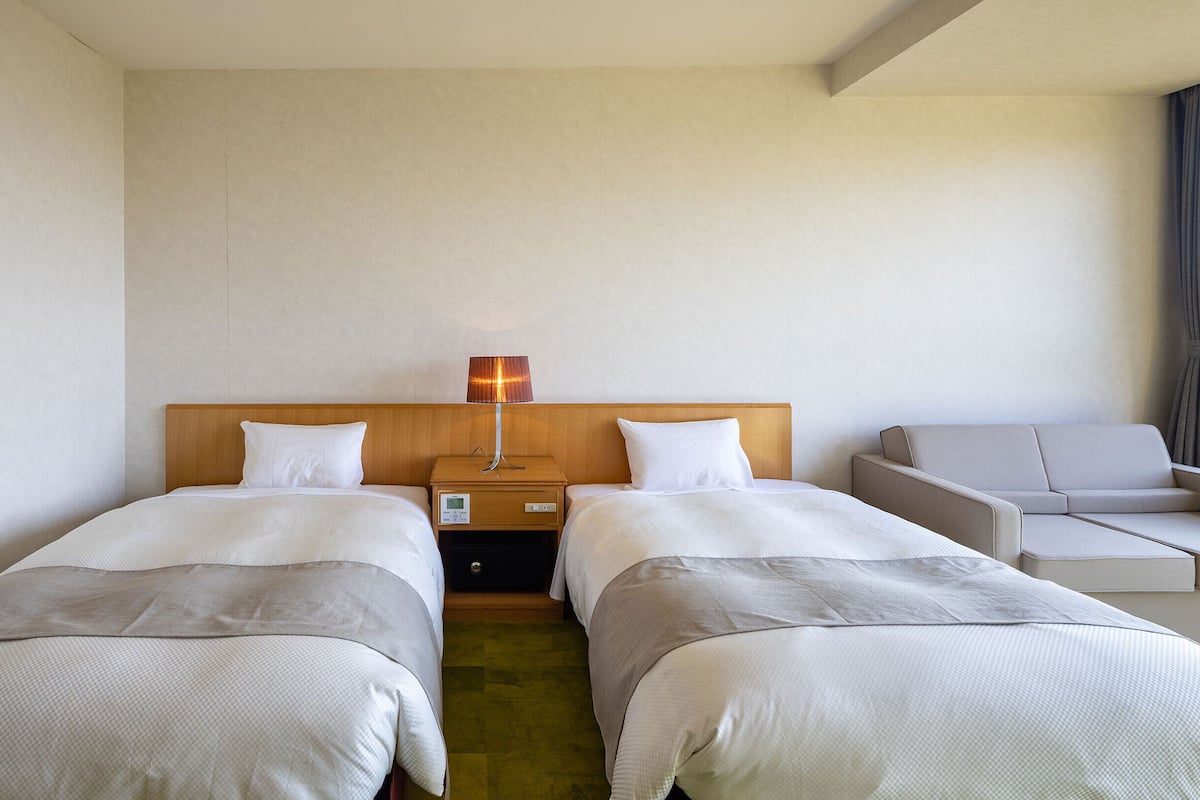 A hotel room with two beds, a bedside table with a lamp, a small table, and a sofa.