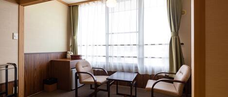 A room with a wooden floor, a small table, two chairs, a window with curtains, and a hanging light fixture.