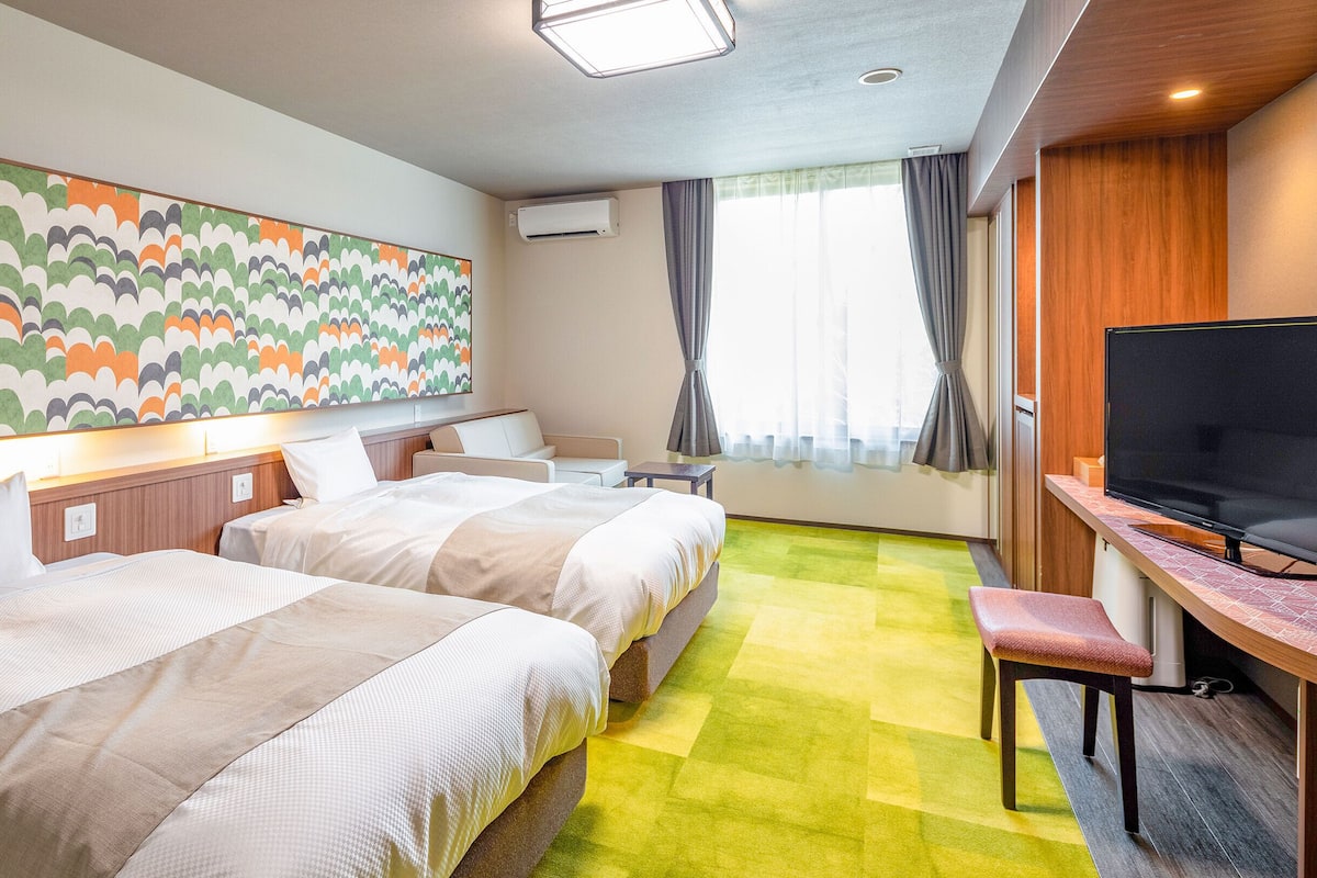 A hotel room with two beds, a TV, a desk, a chair, a large colorful artwork, and a window with curtains.