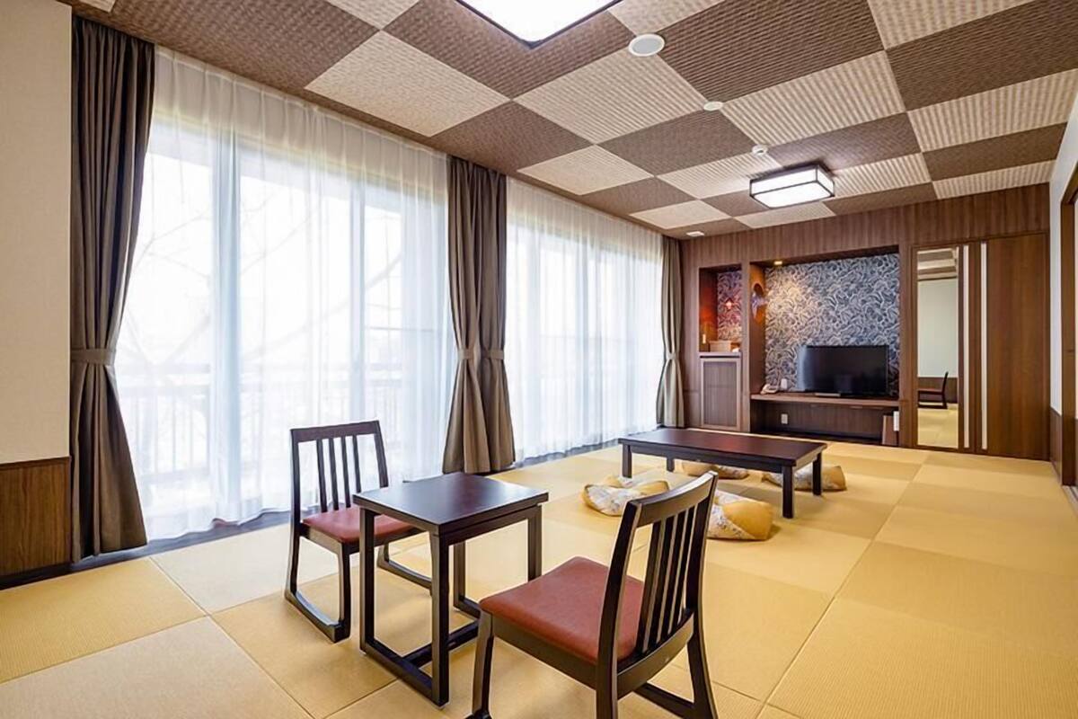 A hotel room with tatami flooring, a traditional Japanese-style dining set, a flat-screen TV, and a decorative wall panel.
