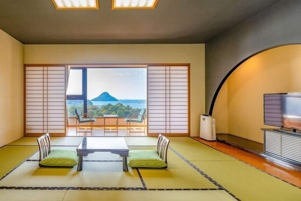 A room with tatami mats, a low table with cushions, a TV, and a view of a mountain and sea.