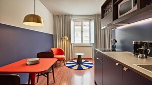 Ein kompaktes Hotelzimmer mit Küchenzeile, einem Essbereich mit rotem Tisch und Stühlen, einem kleinen runden Tisch und Blick auf die Straße durch ein mit Vorhängen drapiertes Fenster.