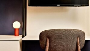 A modern room with a wooden chair, a flat-screen TV mounted on the wall, and a small table with a red lamp. - Studio - Urbanauts Studios Minelli (Trieste)