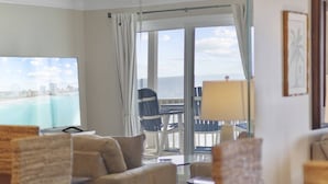Quarto de hotel com uma janela grande que oferece vista para a praia e o horizonte urbano. É possível ver um sofá, uma mesa e uma cadeira.