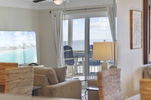 Ein Hotelzimmer mit großem Fenster und Blick auf Strand und Stadtpanorama. Eine Couch, ein Tisch und ein Stuhl sind zu sehen.