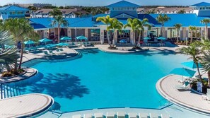 Een zwembadgedeelte van een resort met meerdere ligbedden en parasols, omringd door palmbomen en een groot gebouw met een blauw dak.