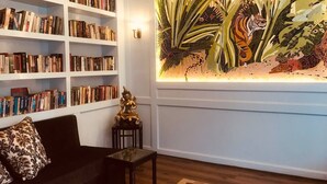 A living room with a bookshelf, a black sofa, a small table with a statue, and a large mural of a tiger and foliage. - Courtyard Pujan Hotel (Kathmandu)