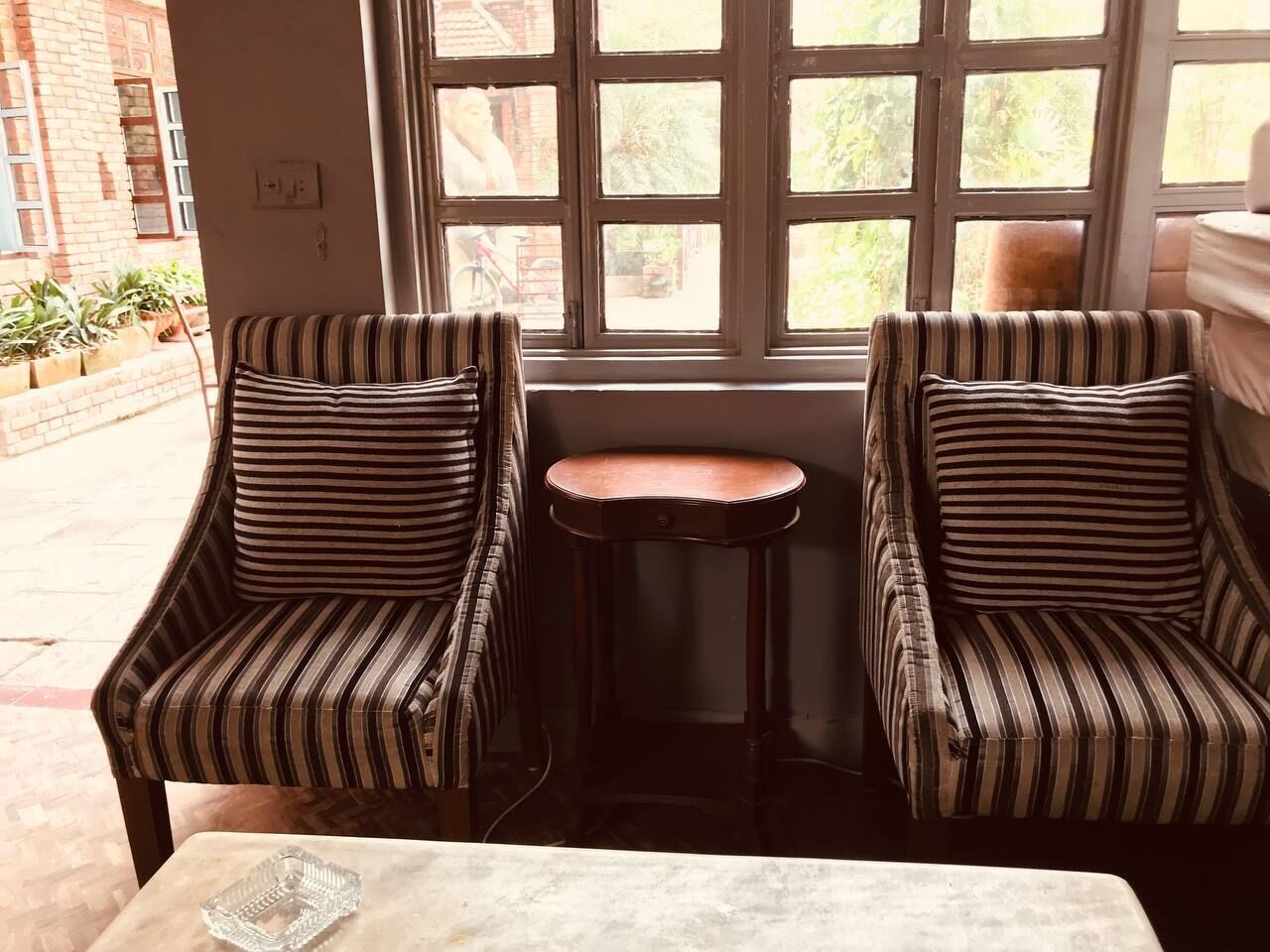A room with two striped armchairs and a small round table, a window with a view of the outside, and a brick wall.