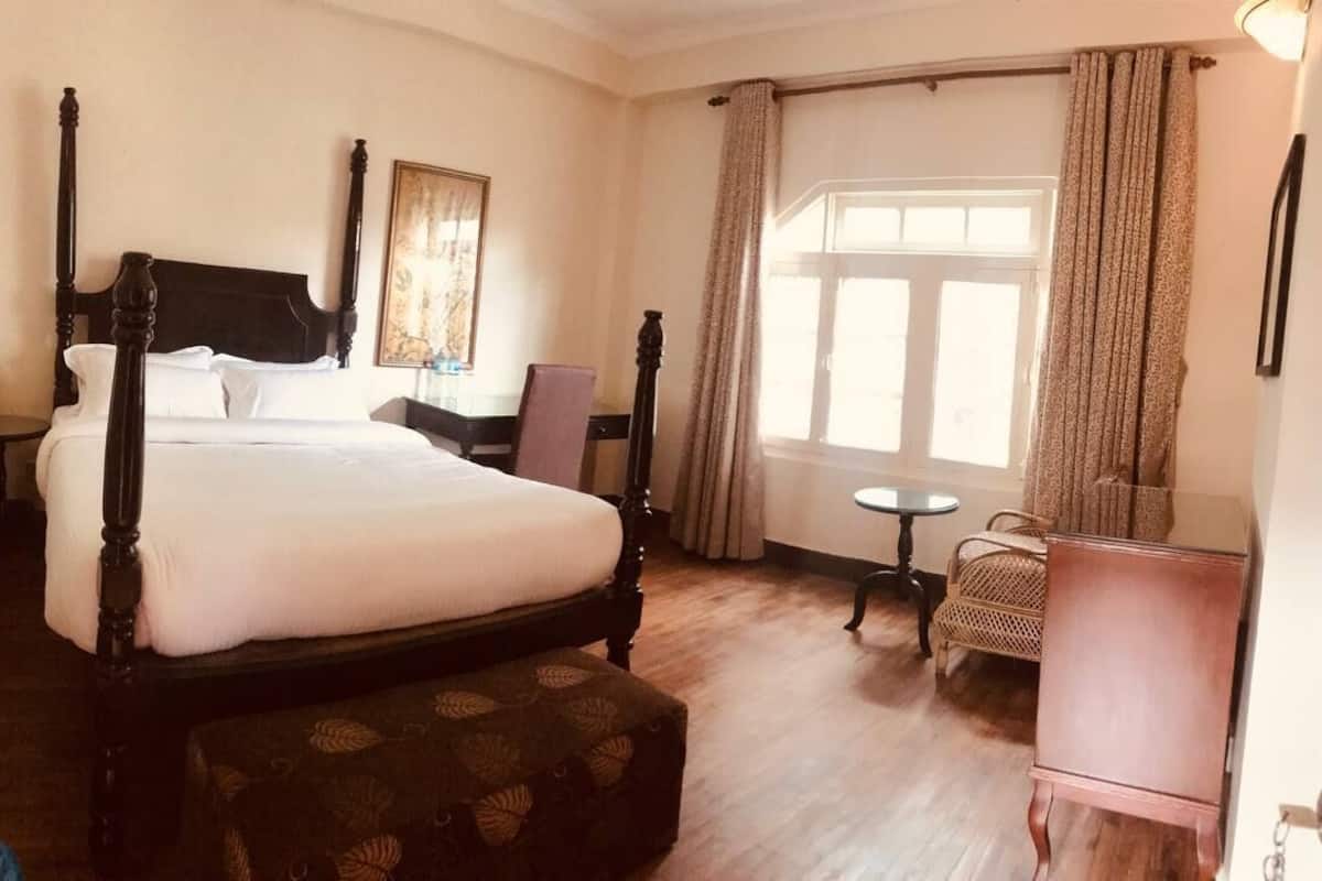 Un dormitorio con una cama con dosel, una mesita redonda pequeña, una silla y un gran ventanal con cortinas.