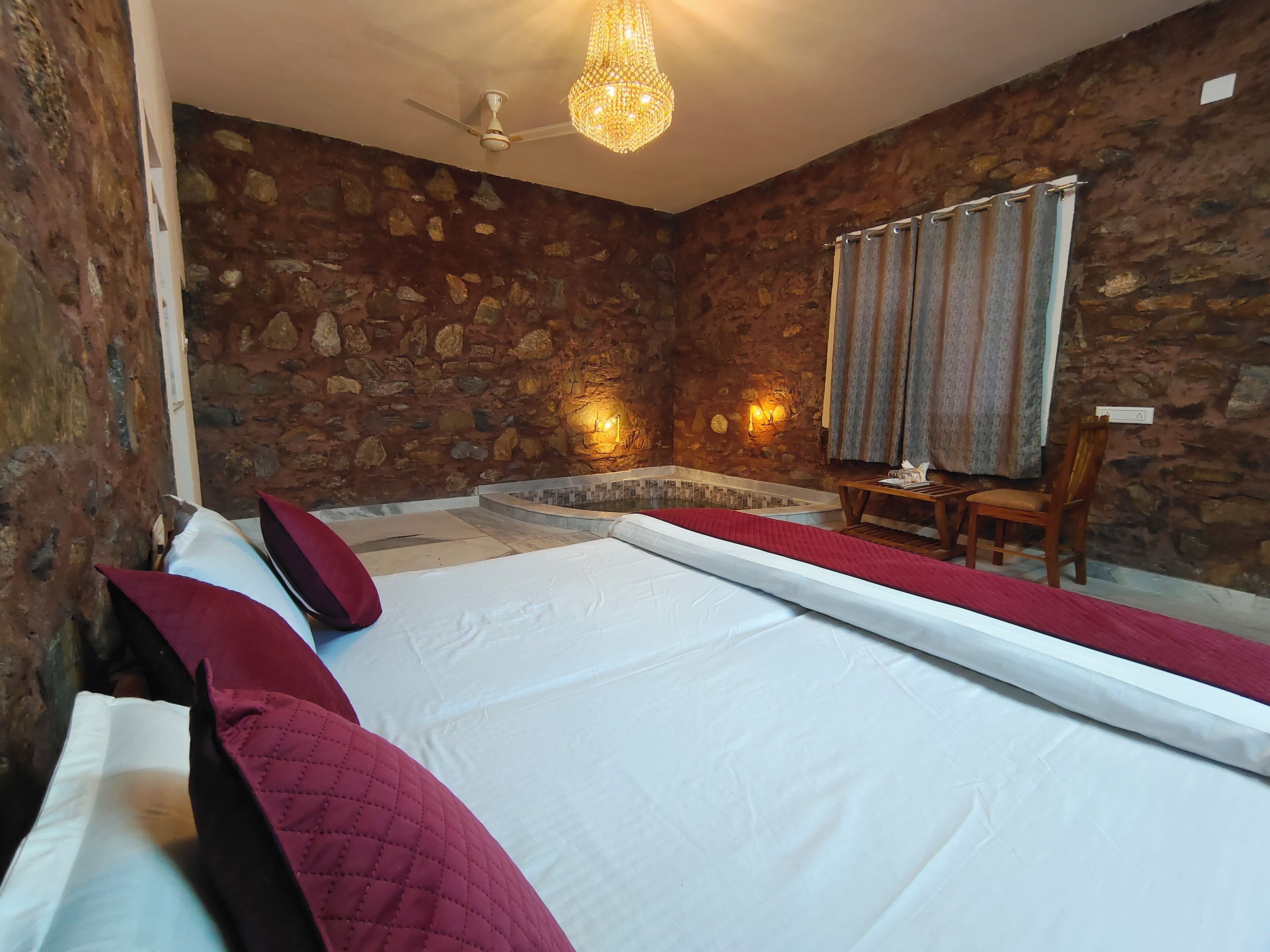 A room with a stone wall, a bed with white linens, a red pillow, a wooden chair, a small table, and a ceiling fan.
