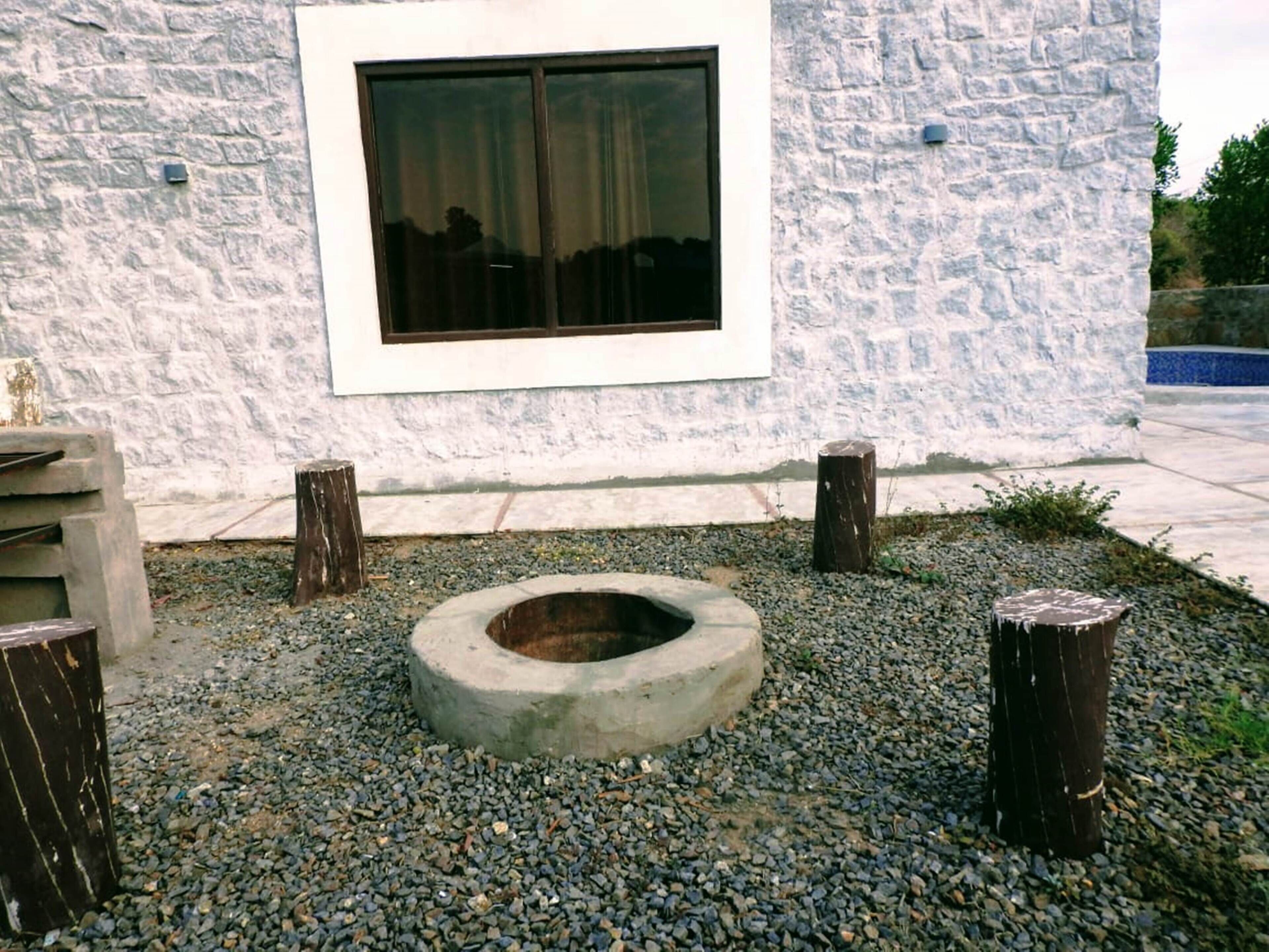 A stone building with a window and a concrete fire pit surrounded by pebbles.