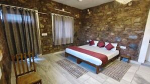 Um quarto com parede de pedra, uma cama com roupa de cama vermelha e branca, um banco de madeira e um banquinho pequeno.