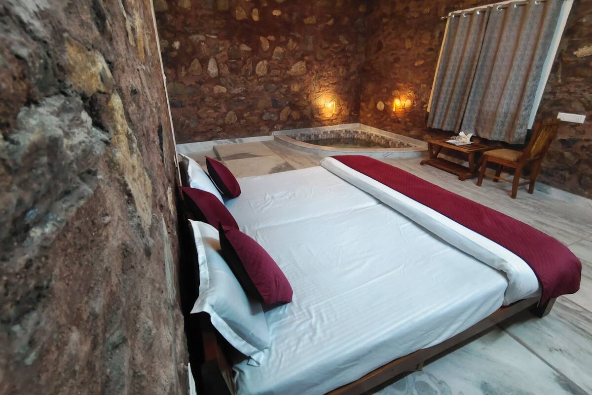 Una habitación con pared de piedra, una cama con ropa blanca y almohadas rojas, un banco de madera y una pequeña zona de estar con una mesa y sillas.