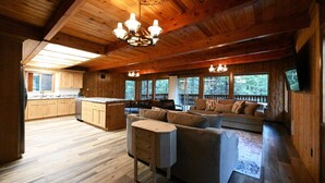 A spacious cabin interior with a wooden floor, a sofa set, a dining area, and large windows offering views of the surrounding forest.