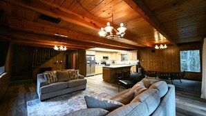 A spacious living area with a wooden ceiling, a kitchen in the background, and a large window providing natural light.