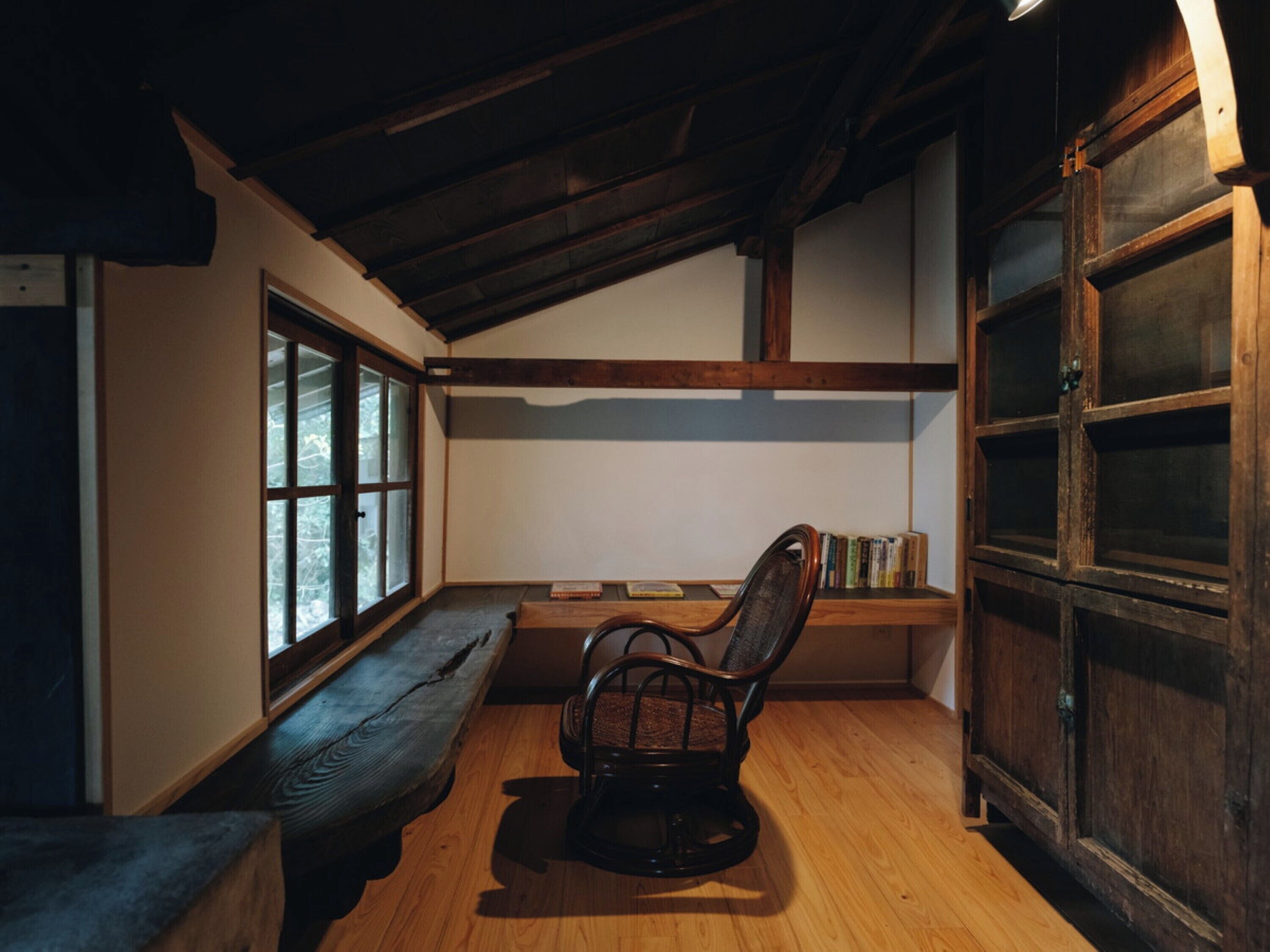 A cozy reading nook with a wooden bench, a chair, and a bookshelf in a room with wooden beams and large windows.