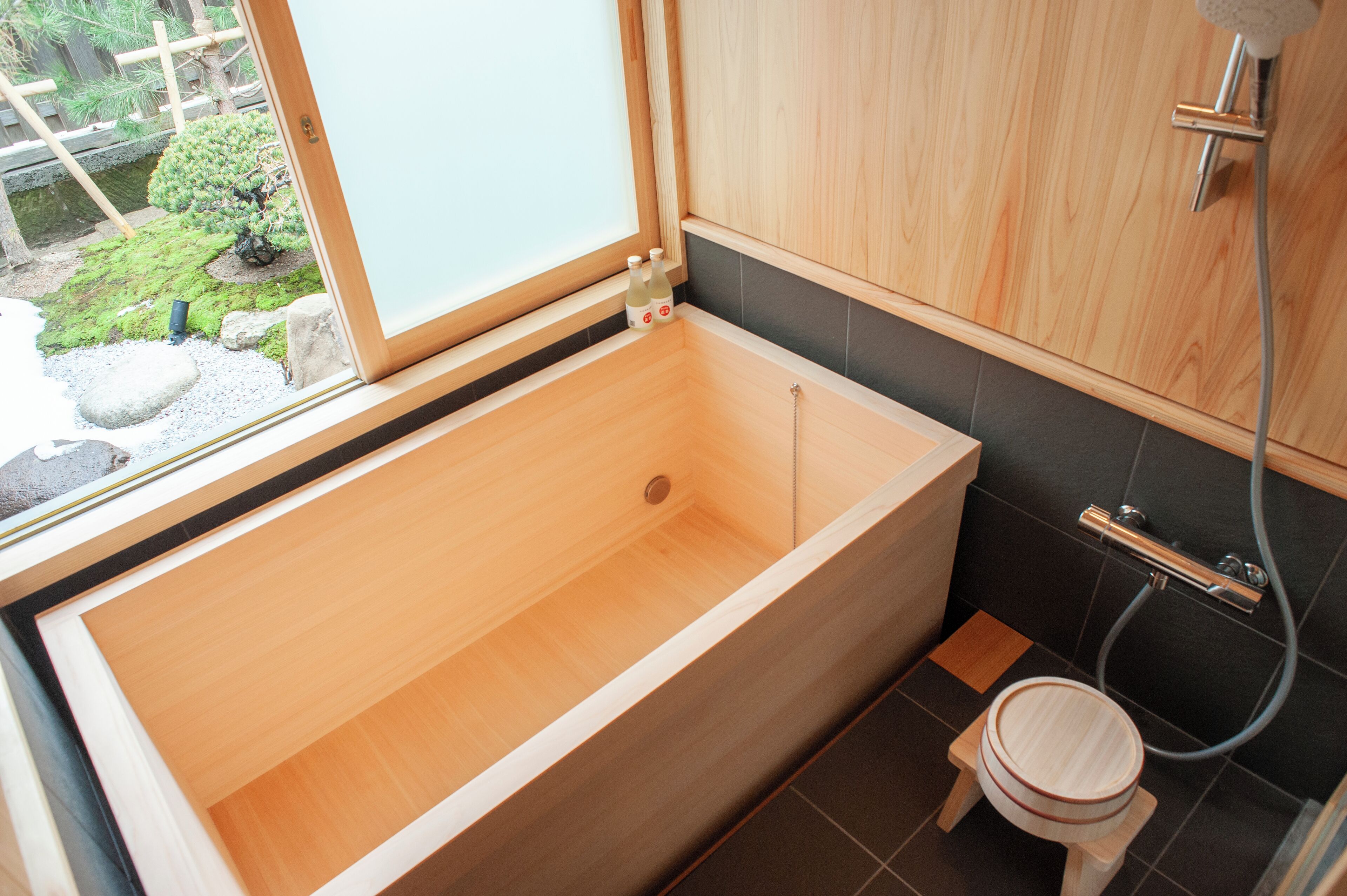 Un baño moderno con una bañera de madera, un taburete pequeño de madera y una ventana con vistas a un jardín.