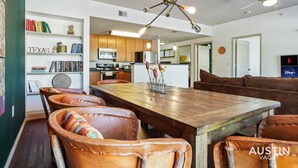 A spacious dining area with a wooden table and leather chairs, a kitchen in the background, and a bookshelf with books and decorative items. - TNC #205 xxxxxxxxxxx (Austin)
