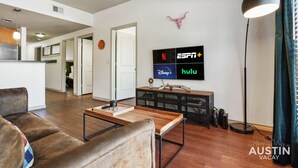 A modern living room with a flat-screen TV displaying streaming services, a wooden coffee table, and a brown leather sofa. - TNC #205 xxxxxxxxxxx (Austin)